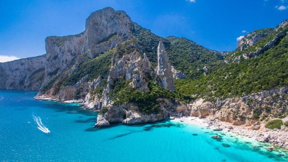 Mediterrane Felsküste mit belebtem Sandstrand in einer kleinen Bucht am türkisblauen Meer unter blauem Himmel.