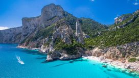 Mediterrane Felsküste mit belebtem Sandstrand in einer kleinen Bucht am türkisblauen Meer unter blauem Himmel.