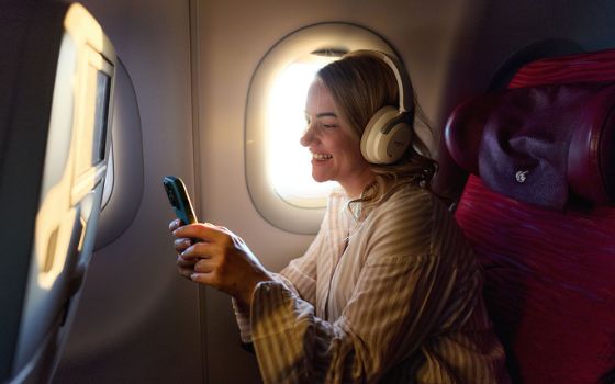 Eine Frau mit Kopfhörern sitzt auf einem Fensterplatz in einem Flugzeug und schaut gut gelaunt auf ein Smartphone in ihren Händen. 