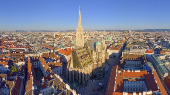 Luftaufnahme der Stadt Wien mit Stephansdom und seinem bunten Dach im Fokus, rundherum Häuserblöcke. 