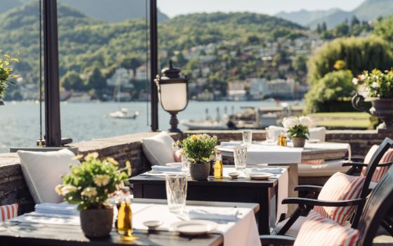 Aussenbereich eines Restaurants, in dem mehrere Tische mit Blumen dekoriert und gedeckt sind, im Hintergrund ein See und grün bewachsene Hügel.