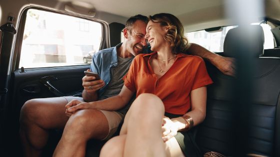 Ein lachendes, sich umarmendes Paar mittleren Alters auf der Rückbank eines Autos, er hält ein Smartphone in der Hand. 