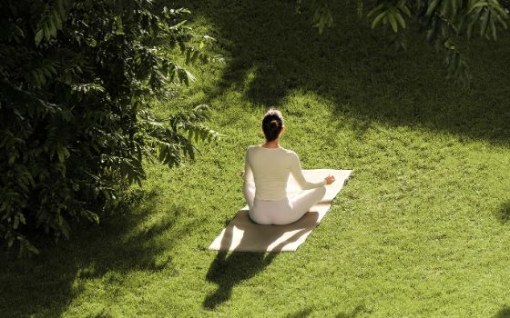 Eine Frau meditiert auf einer weissen Yogamatte in einem grünen Garten.