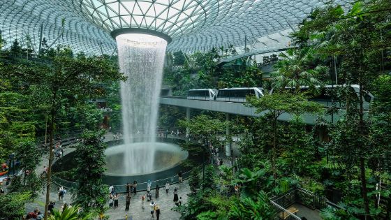 Belebter Flughafenterminal mit Indoor-Garten um einen Wasserfall, der durch eine Glaskuppel fällt und einer Bahn auf einer Hochbahntrasse. 