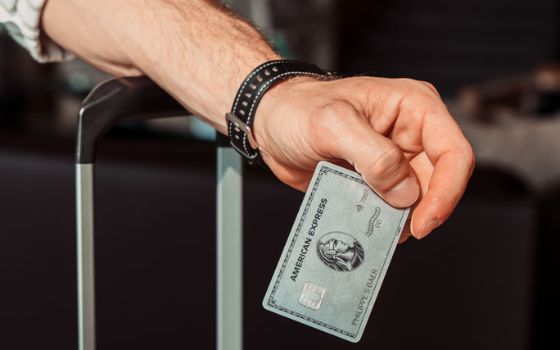 Ein männlicher Arm mit Armbanduhr lehnt auf einem Koffergriff, in der Hand eine silberfarbene Karte mit American Express Logo.