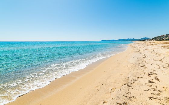 Weitläufiger Sandstrand mit türkisblauem Meer an der Costa Rei im Süden Sardiniens bei klarem Himmel und Blick auf die Hügelkette.