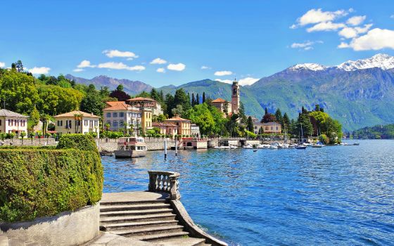 Mediterrane Ortschaft am Ufer eines Sees mit Booten vor Bergpanorama, im Vordergrund eine geschwungene Steintreppe.