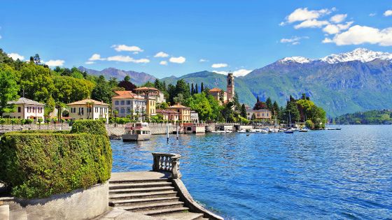 Mediterrane Ortschaft am Ufer eines Sees mit Booten vor Bergpanorama, im Vordergrund eine geschwungene Steintreppe.