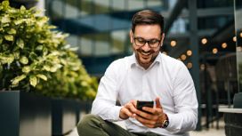 Mann mit Brille und Bart tippt lächelnd auf ein Smartphone.