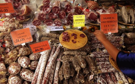 Eine Person greift in die Auslage an einem Marktstand mit Wurstwaren und italienischsprachigen Preisschildern. 