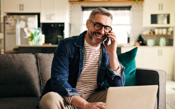 Mann mit grauem Haar und moderner Brille telefoniert und blickt dabei lachend auf einen Laptop.