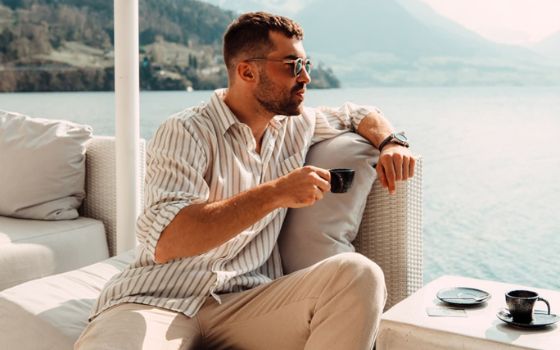Mann sitzt auf einem Sofa mit einem Arm auf der Rückenlehne und hält eine Tasse in der Hand, im Hintergrund ein See