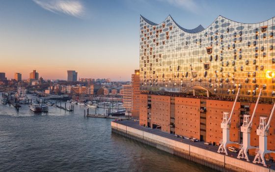 Modernes Operngebäude mit wellenförmigem, gläsernem Korpus auf Backsteinsockel im Hafengebiet Hamburgs bei Sonnenuntergang. 