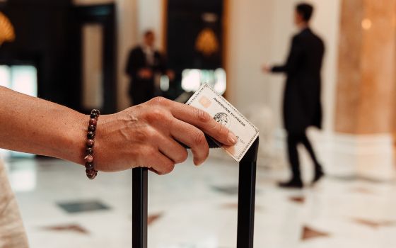 Nahaufnahme einer silbernen Platinum Kreditkarte von American Express in der Hand einer Frau am Griff eines Trolleys in einer edlen Hotellobby.