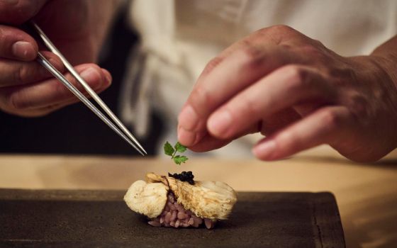 Ein Pilz-Sushi mit schwarzem Kaviar auf einem edlen Steinteller, Hände drapieren ein keines Kräuterblatt auf den Kaviar