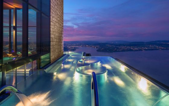 Beleuchteter Infinity-Pool an einem modernen Gebäude mit Blick auf einen See bei Dunkelheit.