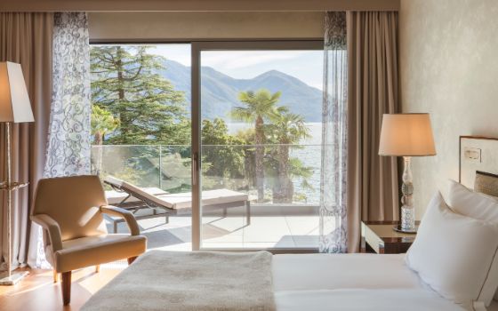 Ein modern eingerichtetes Hotelzimmer in hellen Naturtönen mit Balkon, im Hintergrund grüne Vegetation, Berge und ein See. 