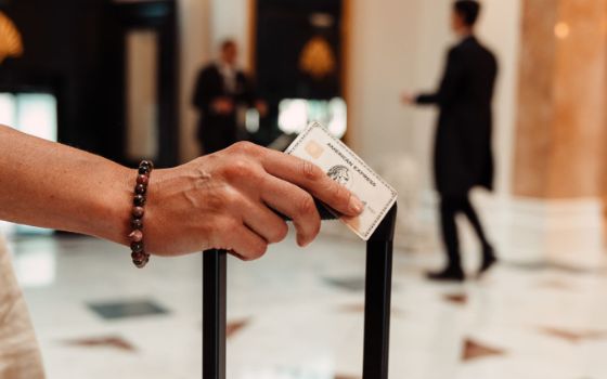 Eine Hand lehnt auf einem Koffergriff und hält eine American Express Karte, im Hintergrund eine Hotellobby.
