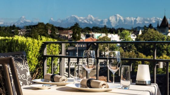 Ein gedeckter Tisch auf einer Terrasse, im Hintergrund sieht man die Berge und eine Stadt mit kleinem Wald.