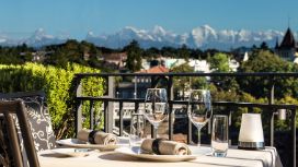 Ein gedeckter Tisch auf einer Terrasse, im Hintergrund sieht man die Berge und eine Stadt mit kleinem Wald.
