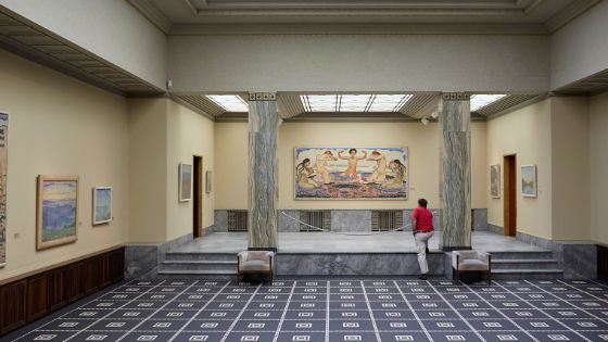 Eine Person vor einem Gemälde im Stil des Symbolismus in einem eleganten Ausstellungsraum mit Marmorsäulen. 
