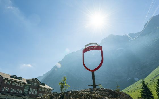 Auf einem kleinen Hügel steht ein rotes Schild mit dem Schriftzug «Grand Tour», im Hintergrund ist eine Berglandschaft bei Sonnenschein zu sehen. 