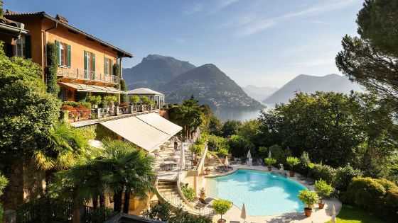 Eine gelbe Villa mit grünen Fensterläden und bepflanztem Balkon hat einen kleinen Swimmingpool, um den Palmen und andere Blumenkübel stehen. Im Hintergrund sieht man einen See und eine Berglandschaft.