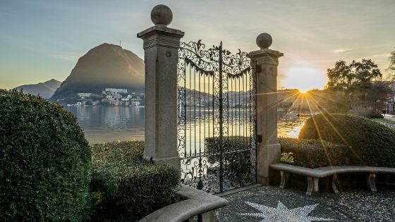 Direkt an einem See liegt ein Tor mit verziertem Eisengitter und einer Buchsbaumhecke, im Hintergrund ragt eine Berglandschaft mit einem kleinen Dorf empor.