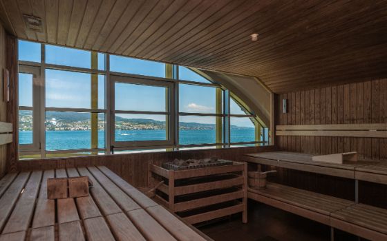 Eine mit Holz verkleidete Sauna mit gegenüberliegenden Bänken und einer Fensterfront, im Hintergrund ein See und grüne Hügel.