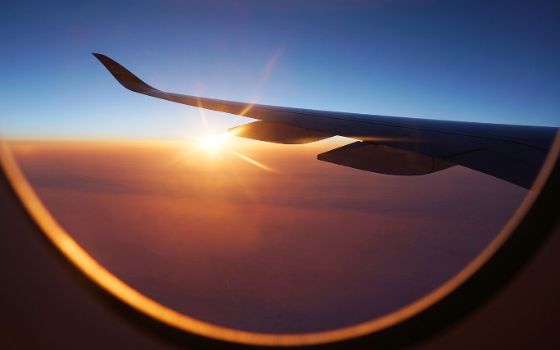 Blick auf die Tragfläche eines Flugzeugs durch ein rundes Flugzeugfenster bei Sonnenuntergang über den Wolken