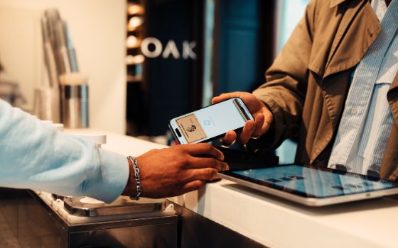 Eine Person in Hemd und Jacke reicht ein Smartphone über einen weissen Tresen, auf dem Display ist eine Kreditkarte von American Express zu sehen. 