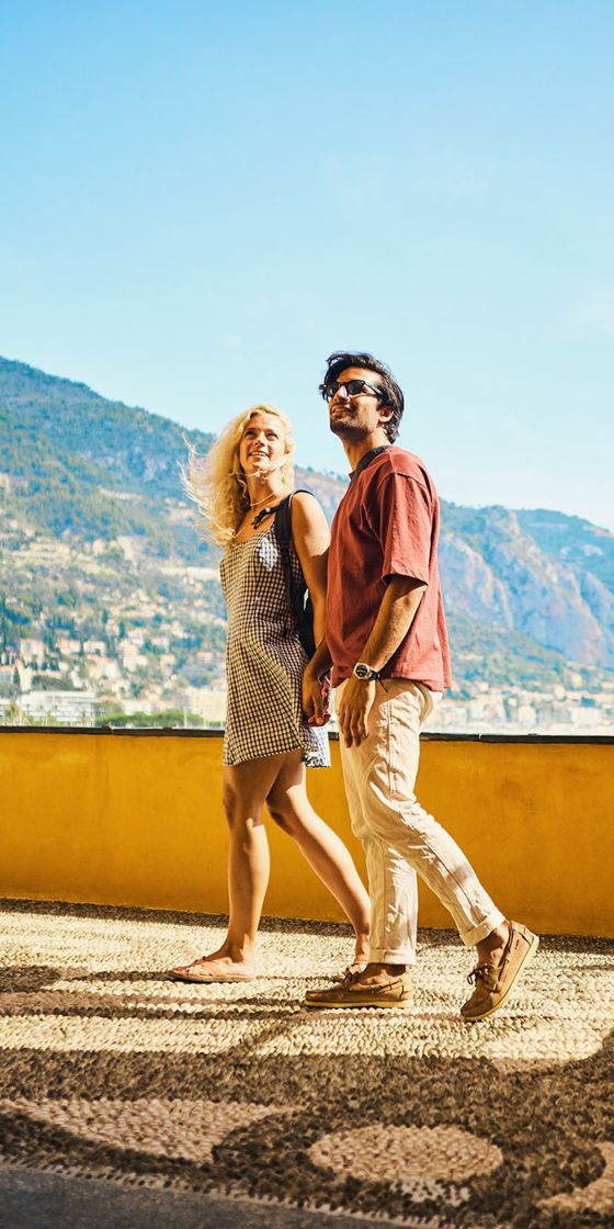 Ein Paar in sommerlicher Kleidung steht händchenhaltend auf einer Plattform mit gelber Mauer vor einer mediterranen Berglandschaft bei Sonnenschein.