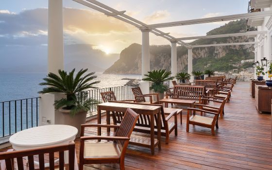 Grosse Hotelterrasse mit Sitzbereich mit zahlreichen Holzmöbelstücken an einer Felsküste bei Sonnenuntergang.