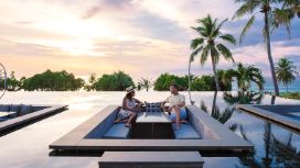 Ein Mann und eine Frau entspannen auf einer Sitzgelegenheit in einer modernen Poollandschaft mit Palmen am Meer bei Sonnenuntergang.