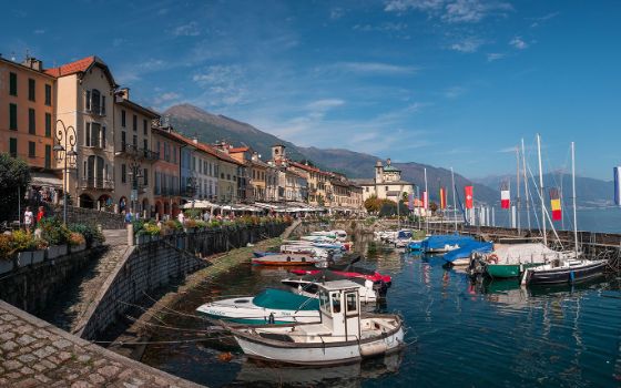 Mediterraner Küstenort mit belebter Promenade und Booten in einem Hafenbecken vor Bergpanorama. 