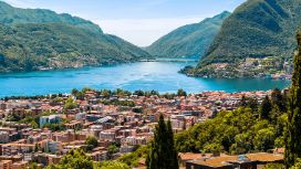 Panorama von Lugano mit Blick auf den Lago di Lugano und die umliegenden Berge bei klarem Wetter, im Vordergrund die Dächer der Stadt.