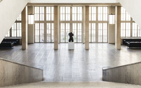 Grosse Steintreppe in einem Museum, die in einen weitläufigen, hellen Raum mit Fensterfront, Steinsäulen und einer Skulptur auf einem Podest führt.