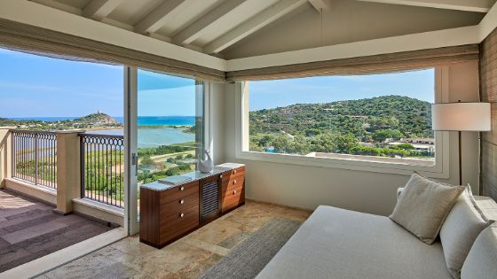 Helle, elegante Hotelsuite mit Terrasse und Blick auf eine grüne, mediterrane Küstenlandschaft unter blauem Himmel.