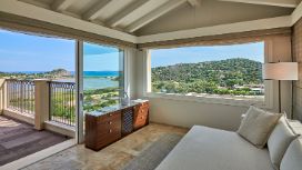 Helle, elegante Hotelsuite mit Terrasse und Blick auf eine grüne, mediterrane Küstenlandschaft unter blauem Himmel.