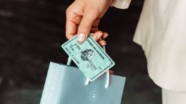 Eine grüne American Express Kreditkarte zusammen mit einer kleinen Papiertragetasche, gehalten von einer weiblichen Hand