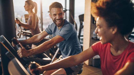 Drei gut gelaunte Personen trainieren nebeneinander an Sportgeräten in einem Fitnessstudio.