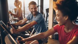 Drei gut gelaunte Personen trainieren nebeneinander an Sportgeräten in einem Fitnessstudio.
