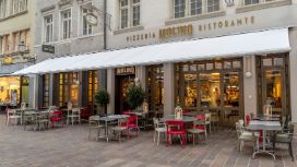 Das Bild zeigt die Aussenansicht des Restaurants mit roten und grauen Stühlen und Tischen, die auf der Strasse stehen.