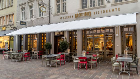 Das Bild zeigt die Aussenansicht des Restaurants mit roten und grauen Stühlen und Tischen, die auf der Strasse stehen.