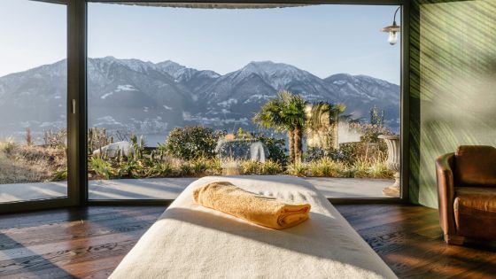 Eine Massageliege steht auf einem dunklen Holzboden. Davor bodentiefe Fenster mit Blick auf Palmen und ein Bergpanorama.