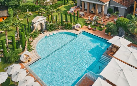 Blick von oben auf einen mediterranen Garten mit Pool, Palmen, Kakteen, Liegen und weissen Sonnenschirmen.  