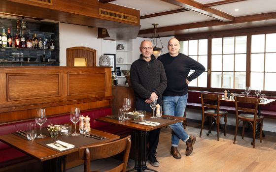 Roman Konrad und Oliver Vaglio stehen nebeneinander zwischen gedeckten Holztischen im Restaurant, im Hintergrund sind grosse Sprossen-Fenster. 