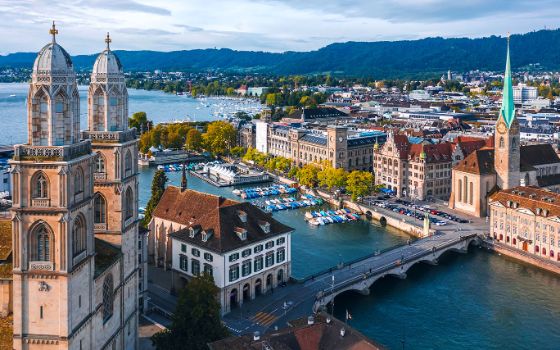 Luftaufnahme Stadtpanorama von Zürich mit zwei Kirchen am Fluss; im Hintergrund ein See, umgeben von grüner Hügellandschaft. 
