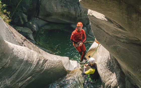 Ein Mann mit Neoprenanzug und Helm ist mit einem Seil gesichert und steht auf einem Bergvorsprung, unten ist ein kleines Wasserloch. 