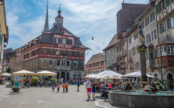 Historischer Marktplatz in Schaffhausen mit bunt bemalten Fassaden, Brunnen und Menschen bei Cafés.